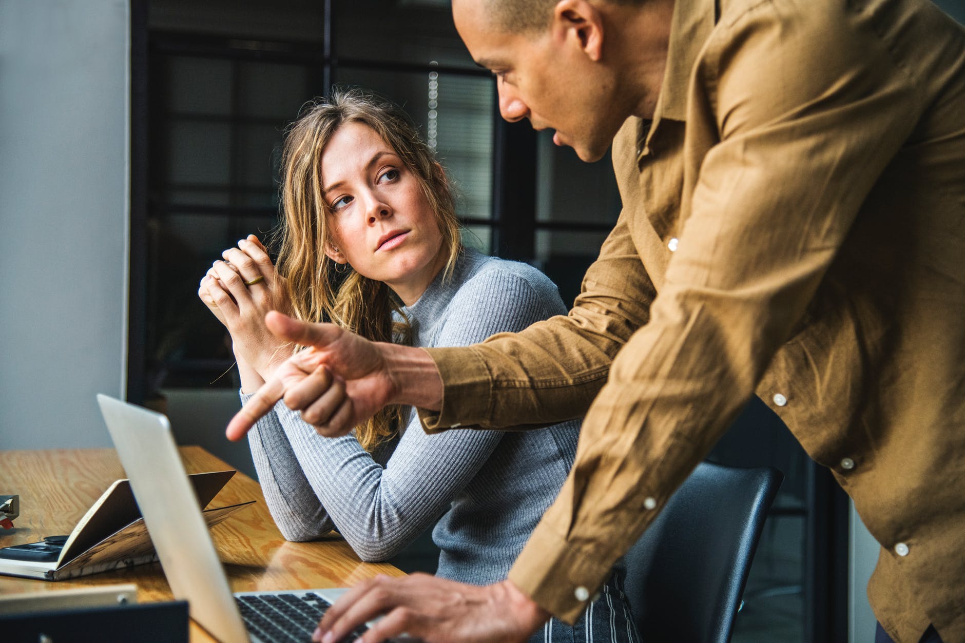 man in brown long sleeved button up shirt standing while using gray laptop computer on brown wooden table beside woman in gray long sleeved shirt sitting